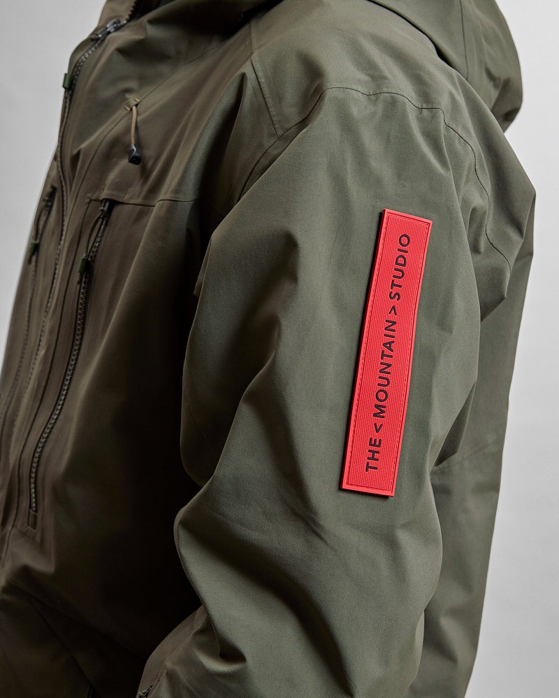 Grüne GORE-TEX Jacke Detailaufnahme einer grünen Z-2 HD GORE-TEX Pro 3L Shell Jacket mit rotem "THE MOUNTAIN STUDIO" Aufnäher am Ärmel. Reissverschlüsse sichtbar.