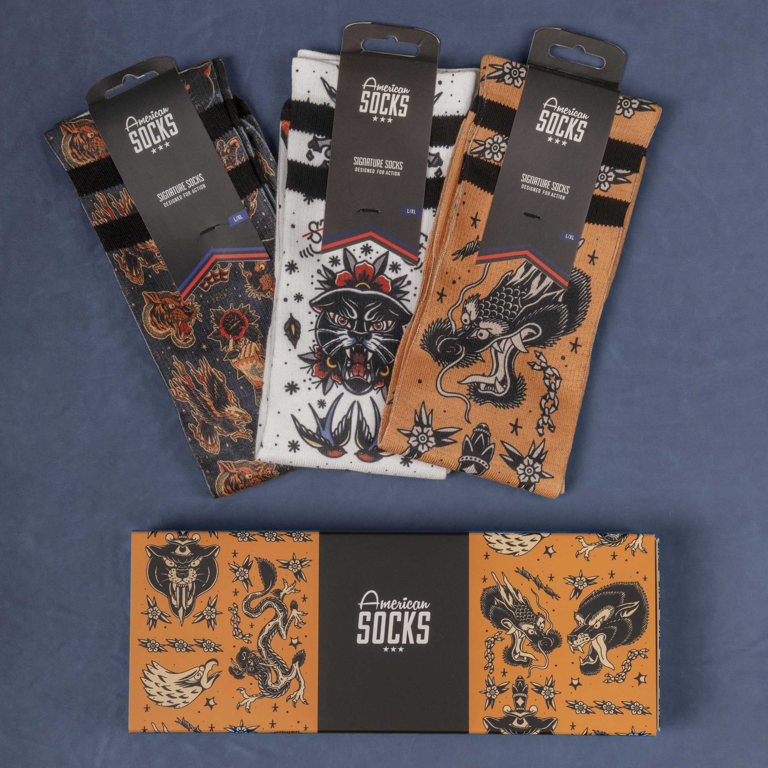Chaussettes de tatouage dans une boîte cadeau Trois American Socks emballés avec des motifs de tatouage et une boîte cadeau assortie sont posés sur un fond bleu.