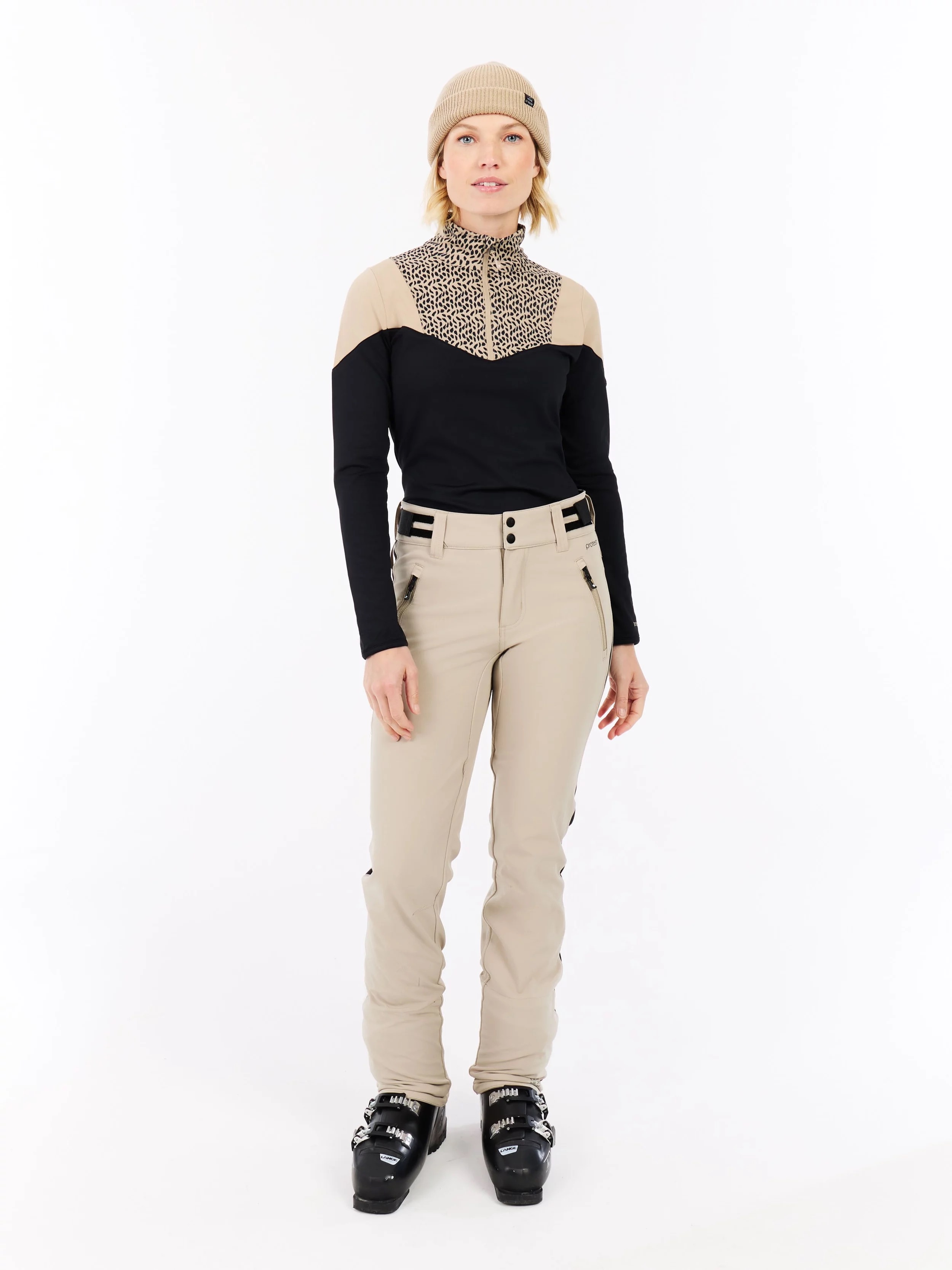 A woman stands in ski clothing in front of a white background. She is wearing a cap, a sweater, pants and ski boots.