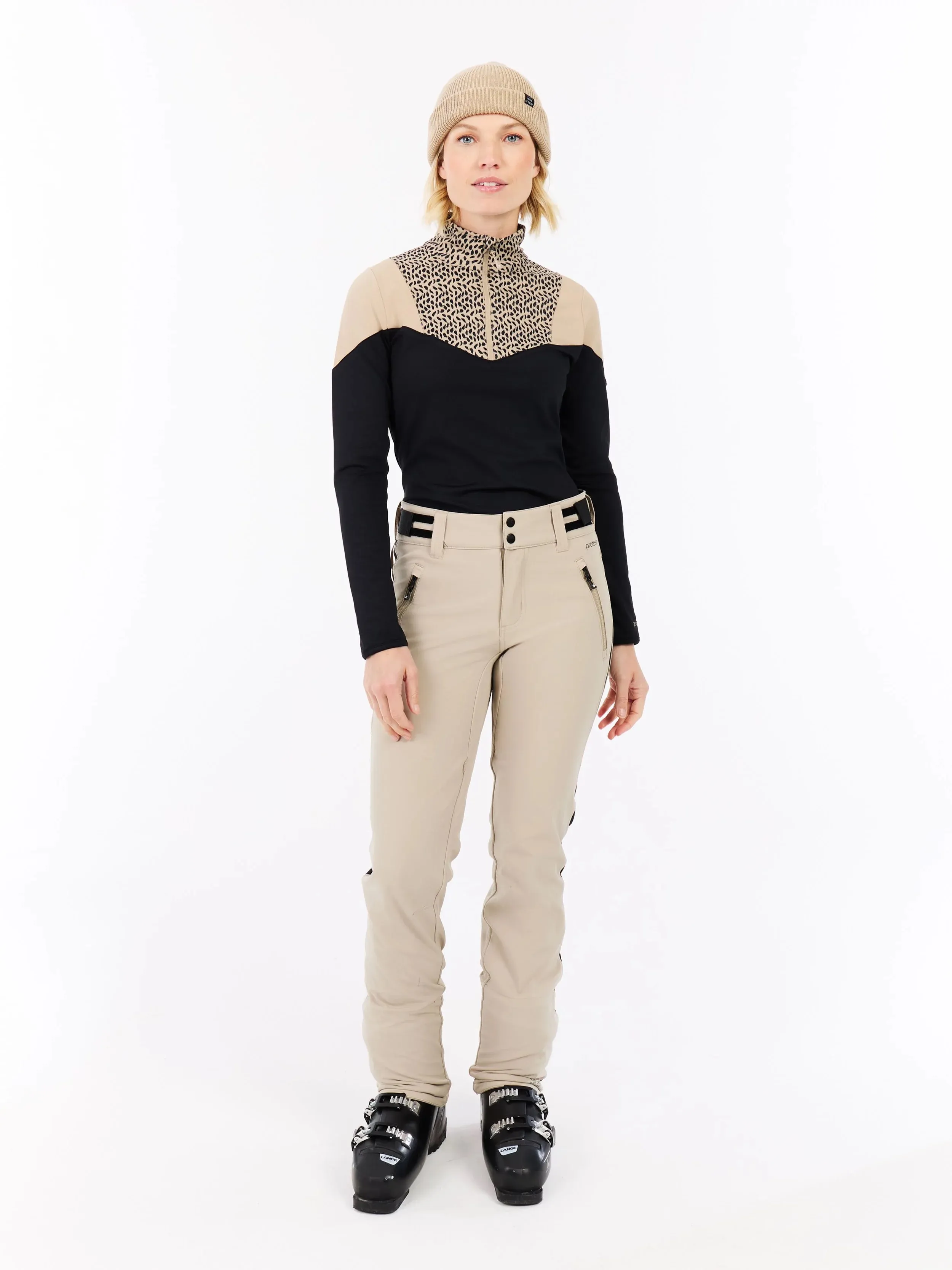 Eine Frau steht in Skikleidung vor einem weißen Hintergrund. Sie trägt eine Mütze, einen Pullover, eine Hose und Skischuhe.