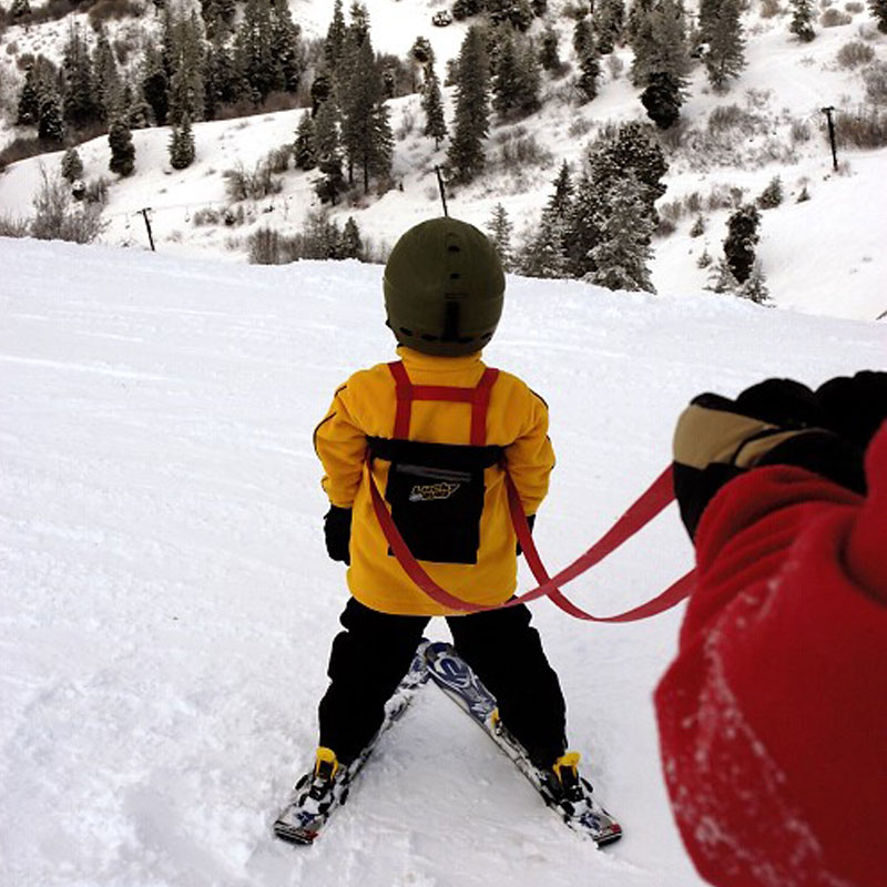 Kind mit Ski Trainer Geschirr und Leine auf Ski, Piste und Bäume im Hintergrund.