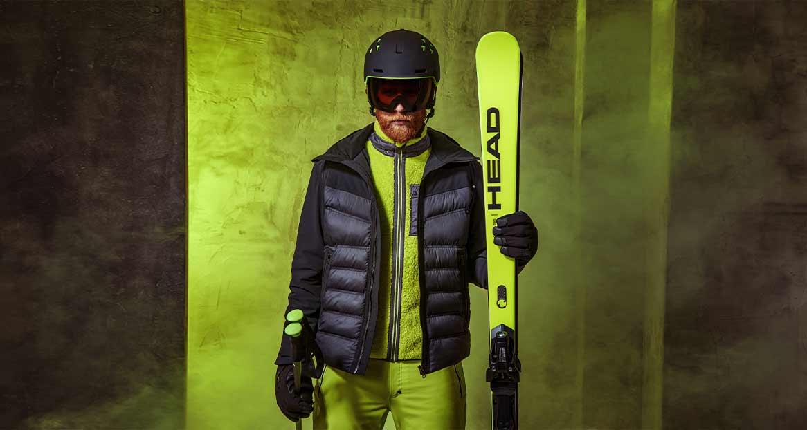 Ein Mann in Skikleidung mit Helm und Brille hält Skier und Stöcke vor einem strukturierten, zweifarbigen Hintergrund.

