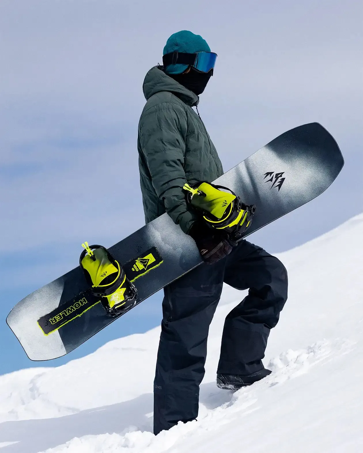 Uno snowboarder con giacca e berretto verde porta sulla neve uno snowboard con attacchi gialli.