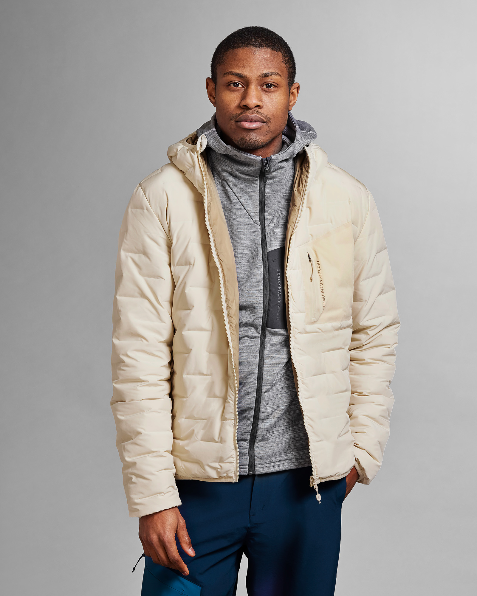 Sandfarbene D-3L Jacke Mann in Sandfarbener D-3L Reversible Light Hood Jacket mit Kapuze, darunter grauer Hoodie und dunkelblaue Hose.