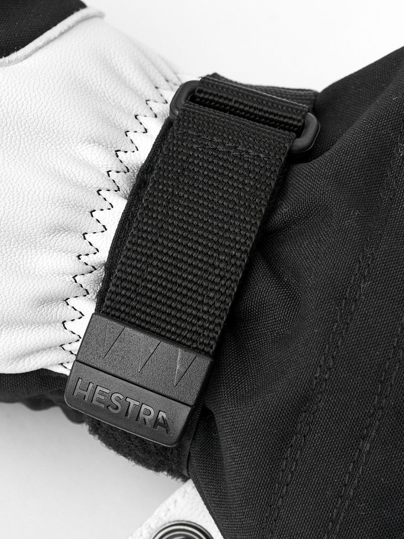 Detailaufnahme eines schwarzen Hestra Skihandschuhs mit weißem Lederteil und einem schwarzen Gurt mit dem Hestra-Logo.