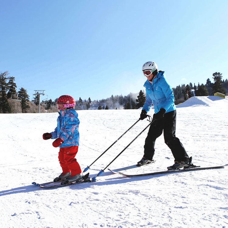 Ein Erwachsener bringt einem Kind auf einer sonnigen Skipiste das Skifahren bei. Er nutzt den Hookease Ski Trainer, um die Skier des Kindes über seine Skistöcke zu steuern. Das Kind trägt roten Helm und bunte Jacke.