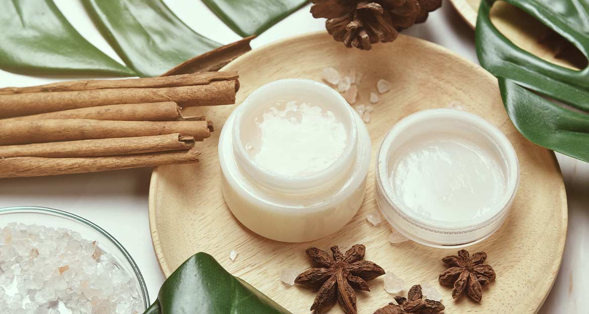Sur une assiette en bois sont disposés deux pots de crème ouverts et remplis, des bâtons de cannelle, de l'anis étoilé, une pomme de pin et une feuille verte.