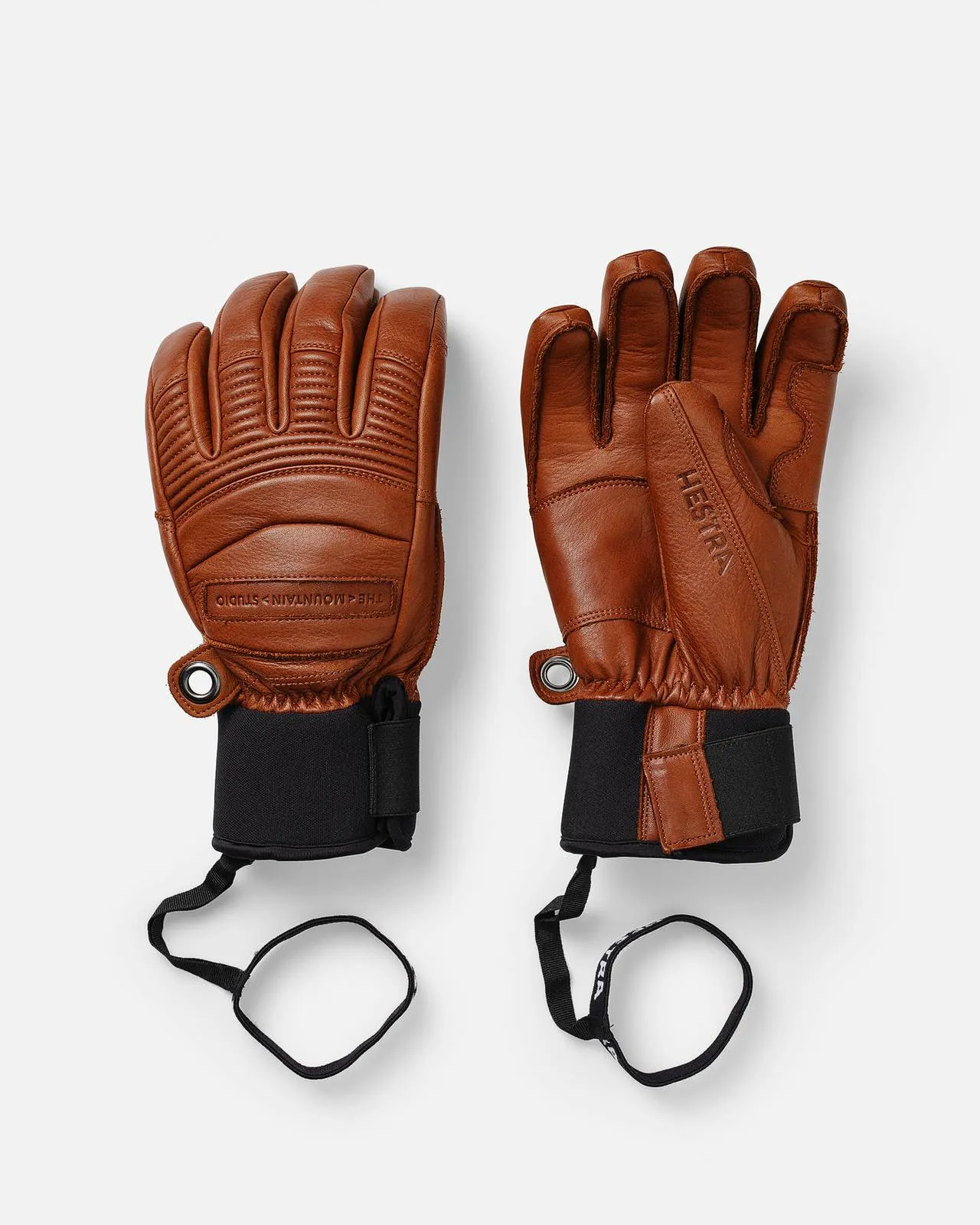 Two brown Hestra leather gloves with black wrist bands on a white background.