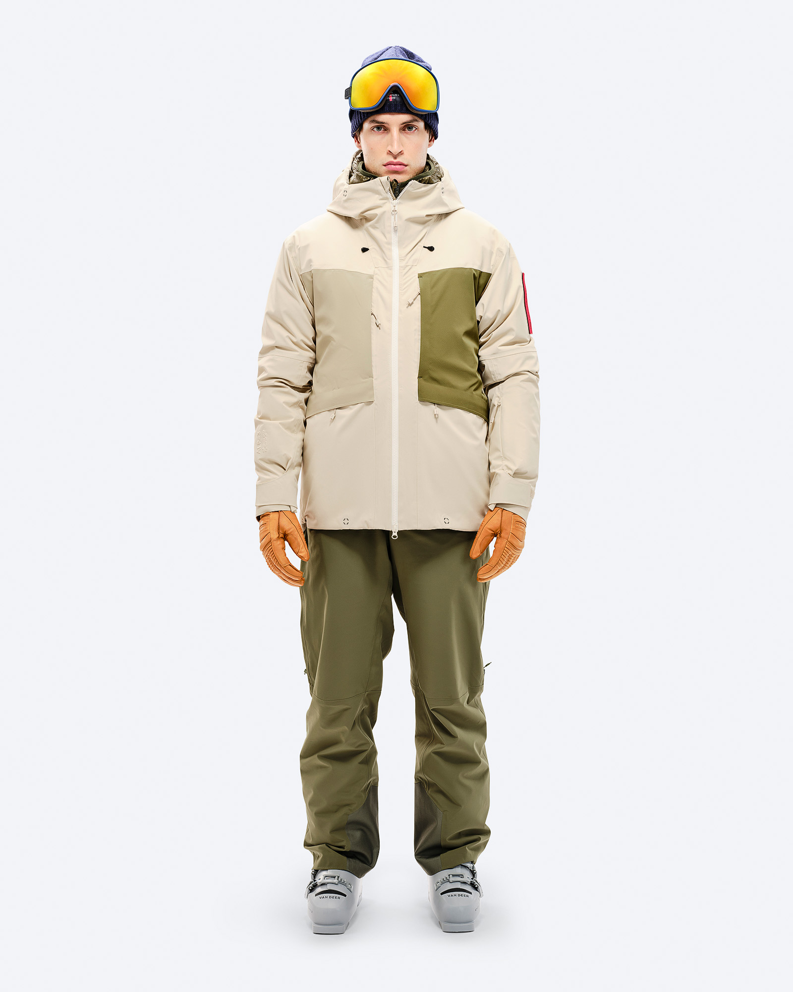 Beige 3L Hybrid Jacket Person in beiger 3L Hybrid Shell Jacket, grüner Hose, Handschuhen und Skibrille vor neutralem Hintergrund.