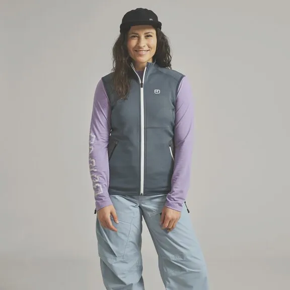Una donna con berretto nero, top viola e gilet grigio sorride su uno sfondo grigio. Indossa pantaloni blu chiaro.