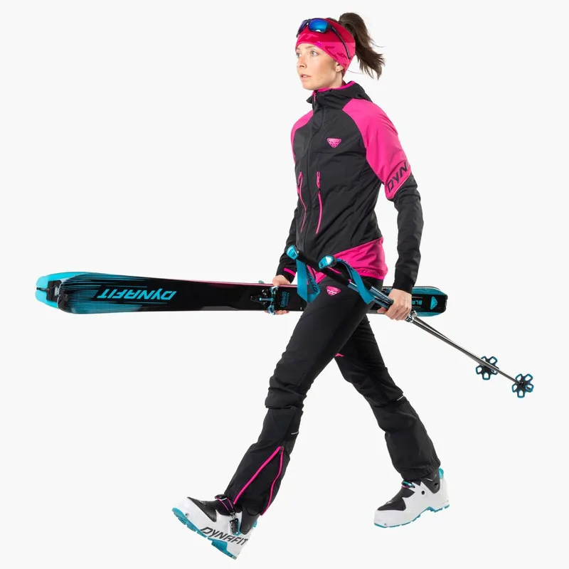 Frau in Skikleidung mit Blacklight 88 Ski unter dem Arm und Skistöcken in der Hand, geht vorwärts.
