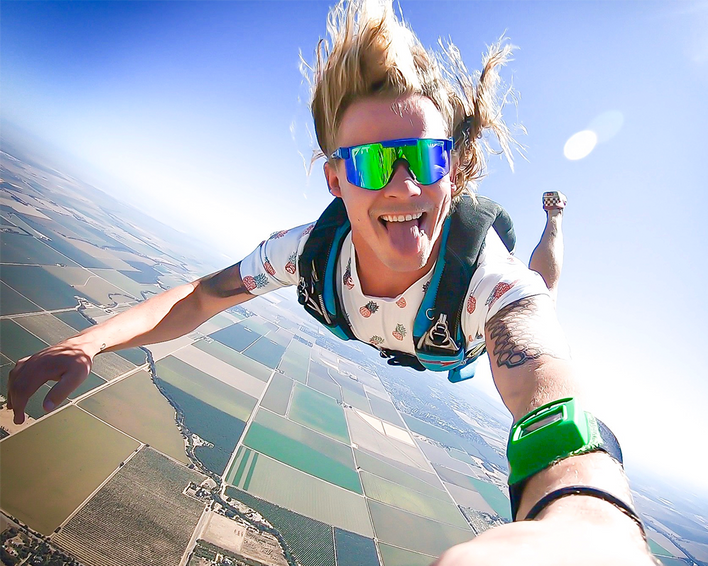 Un homme en chute libre avec des lunettes polarisées bleu-vert qui tire la langue. Des champs sont visibles en arrière-plan.