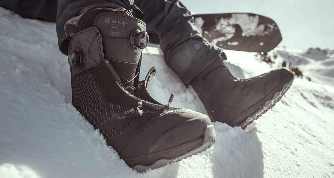 Chaussures de snowboard noires de la marque Nidecker dans la neige. En arrière-plan, on peut voir un snowboard et un paysage de montagne enneigé.