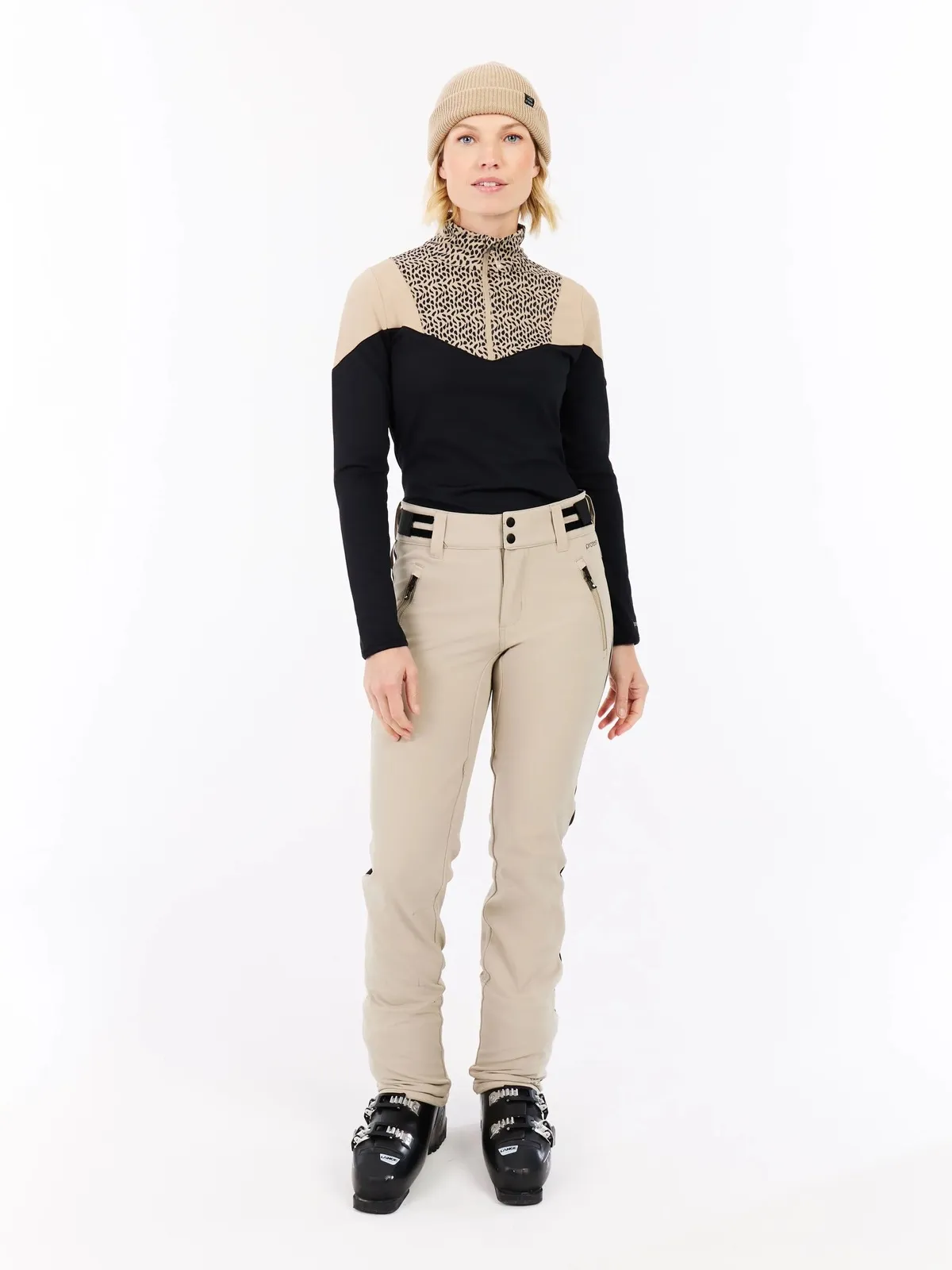 Eine Frau steht in Skikleidung vor einem weißen Hintergrund. Sie trägt eine Mütze, einen Pullover, eine Hose und Skischuhe.