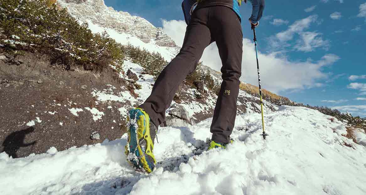 Wanderer mit Schuhkrallen und Stock im Schnee; Berggipfel und blauer Himmel im Hintergrund.