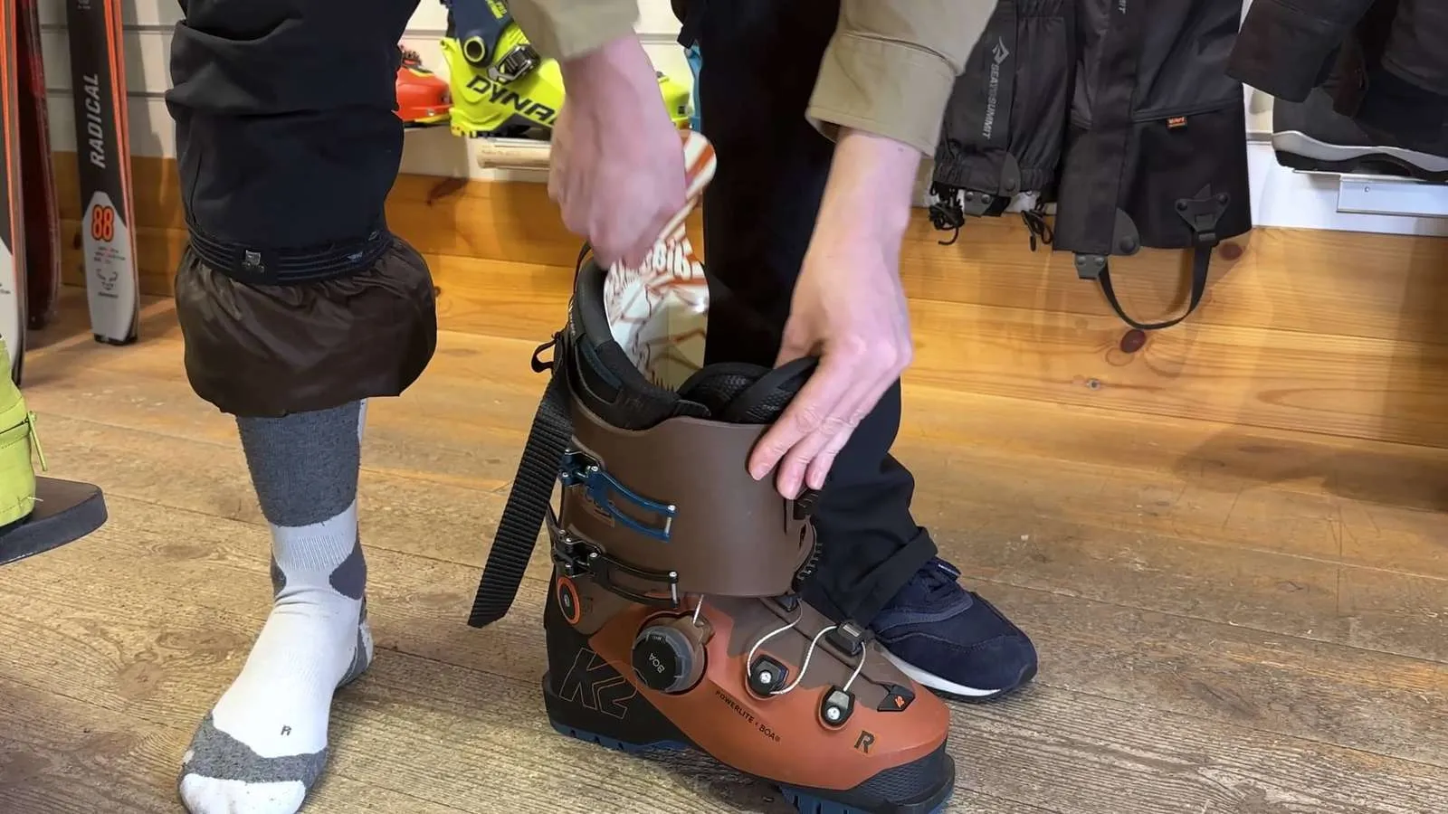 Person puts on a ski boot. Brown K2 Powerlite ski boot with Boa system is put on. Socks and gaiters are visible.