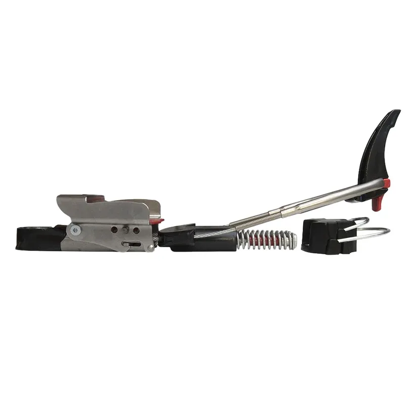 Fixation de ski de randonnée argentée sur fond blanc : mâchoire avant avec touches rouges, câble et ressort rouges, talonnière noire, bloc noir dessous.