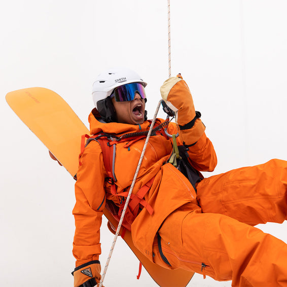 Person mit Snowboard, orangefarbener Kleidung, weißem Helm und THE SHABOOMS SYNTHESIZER Sonnenbrille an einem Seil hängend.
