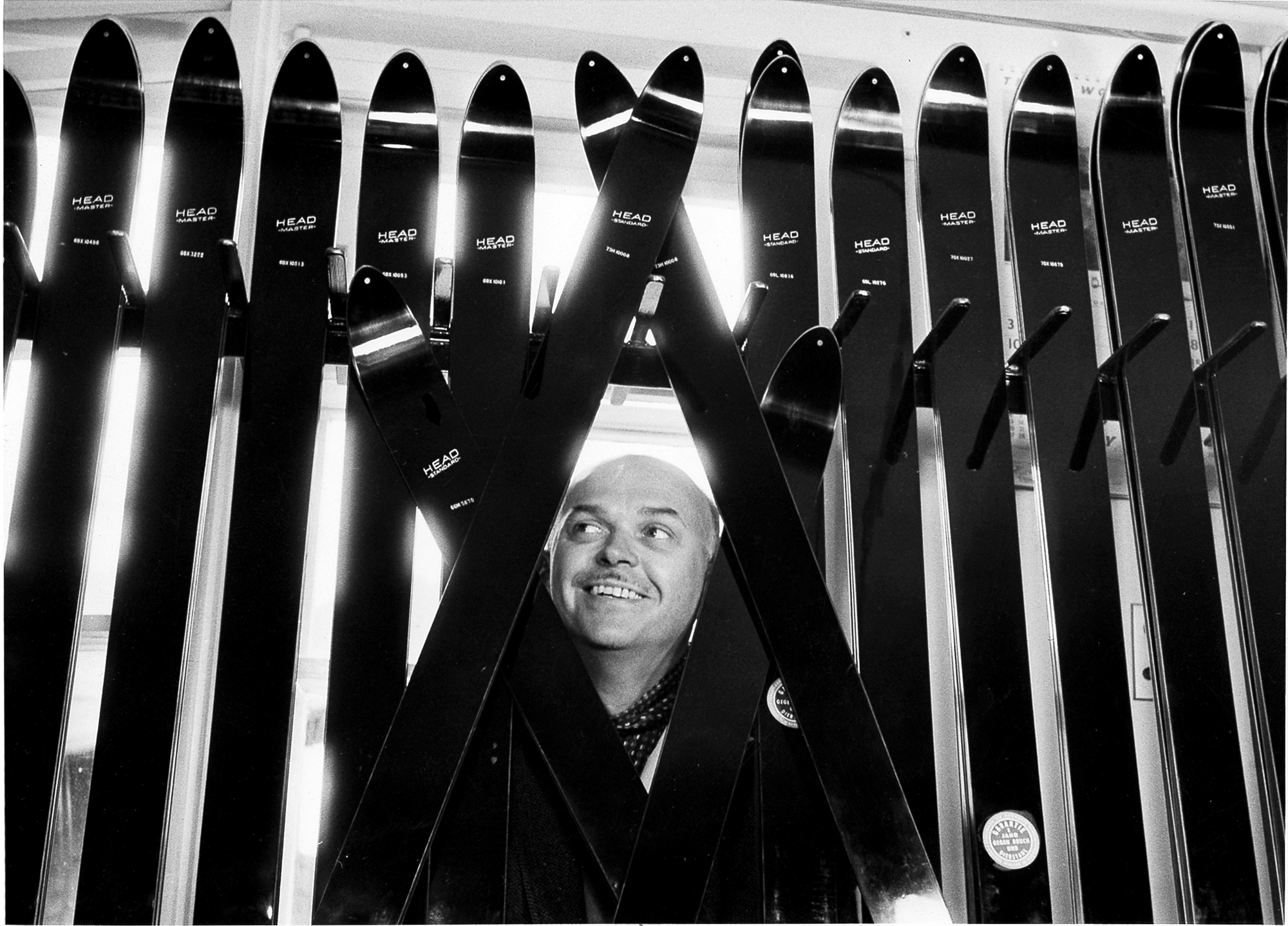 Lächelnder Skifahrer Schwarzweiß-Porträt eines Mannes, der durch gekreuzte Skier hindurchschaut und lächelt. Im Hintergrund sind weitere Skier zu sehen.