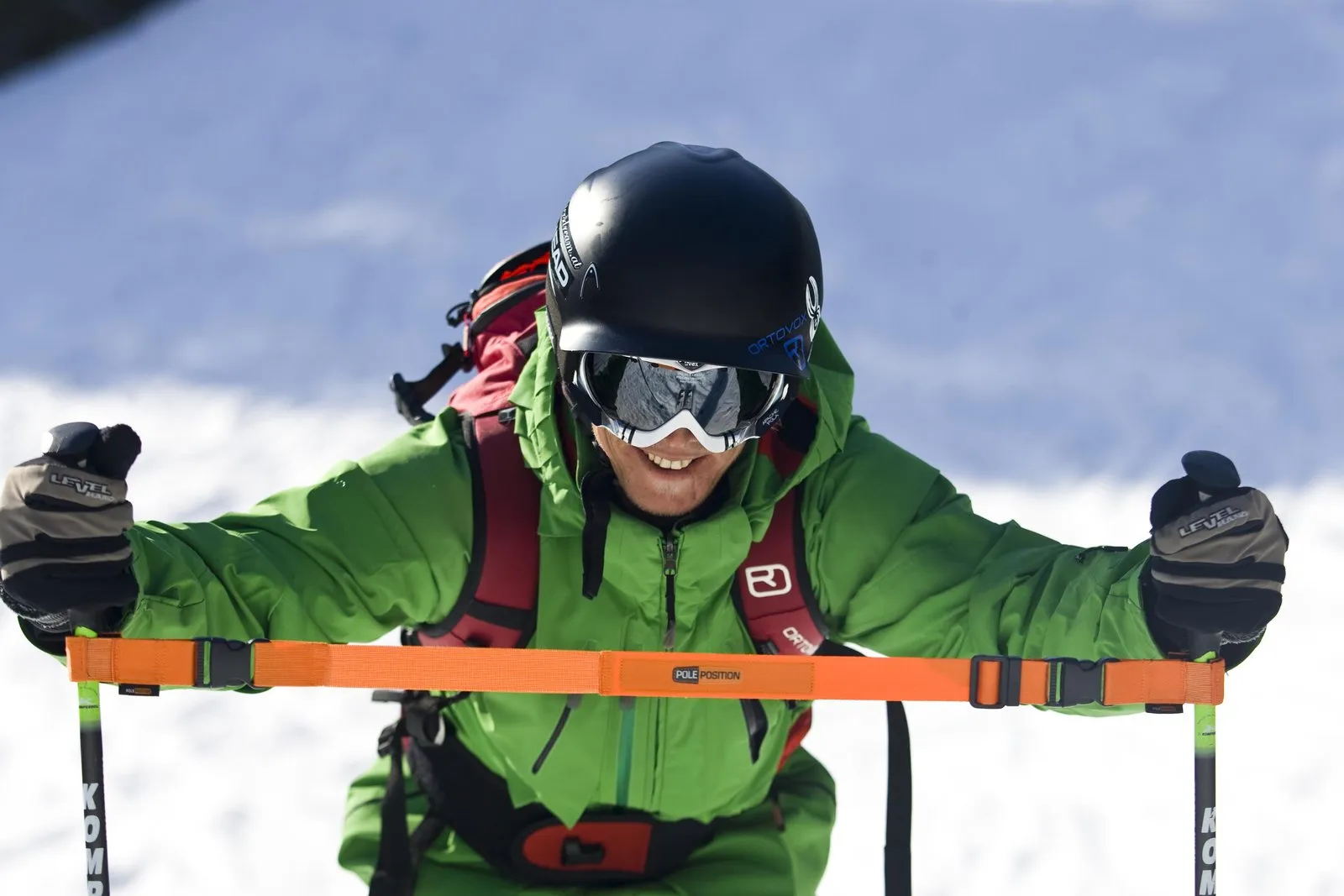 Skier with helmet, ski goggles and green suit holding orange training strap with the lettering "Pole Position".