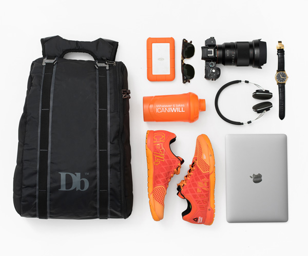 Douchbags The Base 15L Rucksack mit Laptop, Kamera, Kopfhörer und Turnschuhen auf weißem Hintergrund.