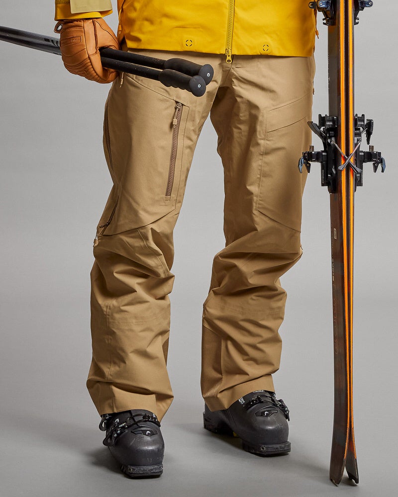 Person in sandfarbener GORE-TEX Pro 3L Shell Pant mit Skiausrüstung. Skistöcke in der Hand, Ski an der Seite.
