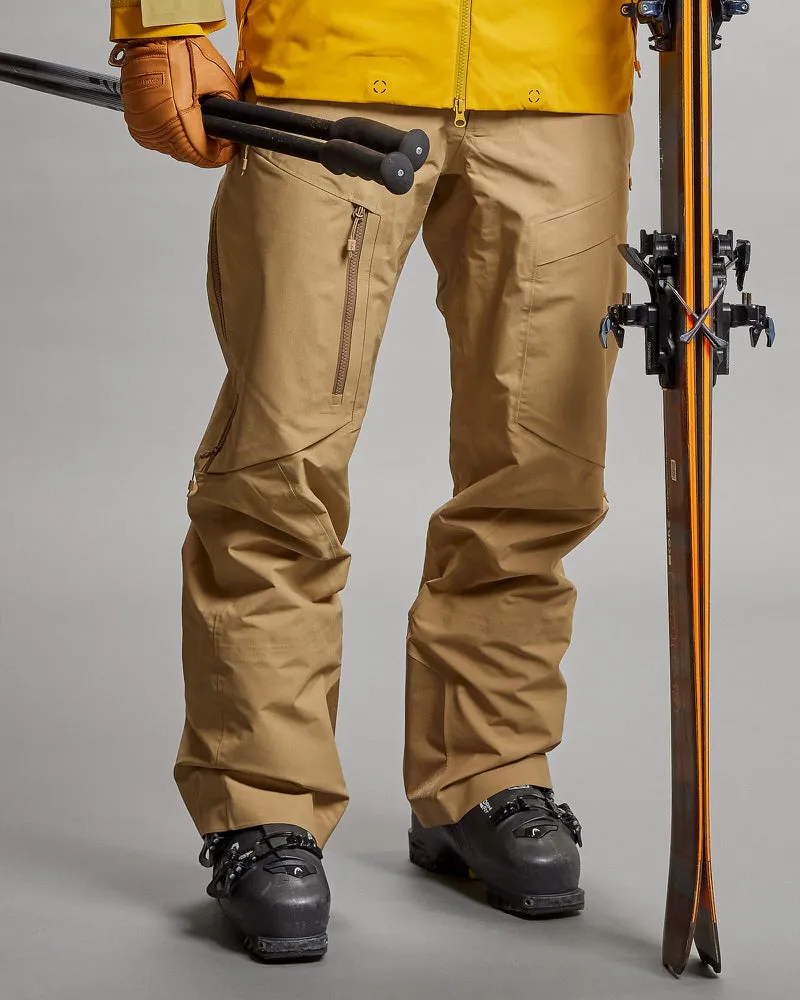 Person in sandfarbener GORE-TEX Pro 3L Shell Pant mit Skiausrüstung. Skistöcke in der Hand, Ski an der Seite.
