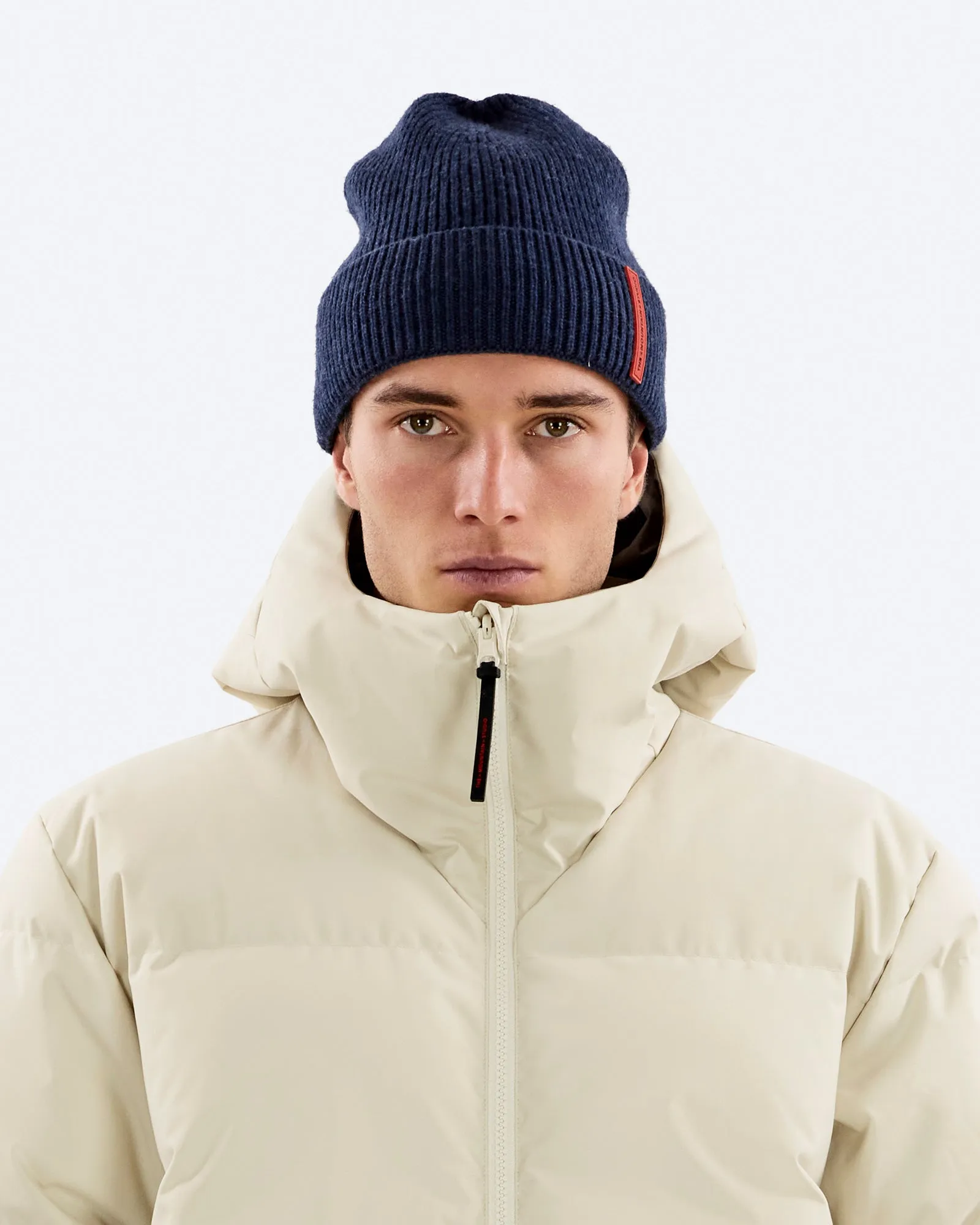 Ritratto di un uomo con berretto blu e piumino beige davanti a uno sfondo bianco.