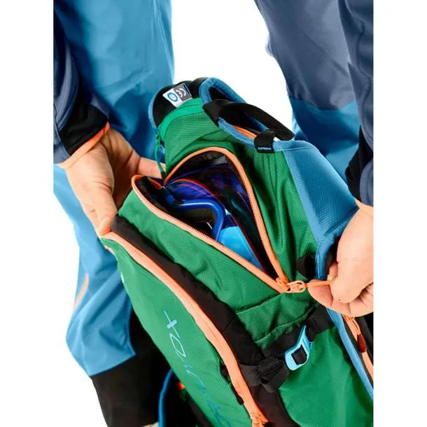 Detailansicht eines grünen Ortovox Free Rider 24 Rucksacks, der von Händen geöffnet wird und eine Skibrille zeigt.