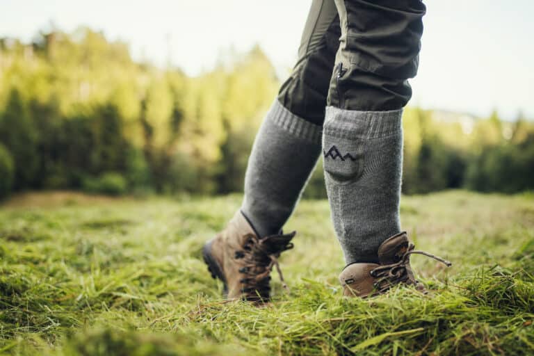Graue Heizsocken V2 Beine mit grauen Everyday Heizsocken V2 und Wanderschuhen im Gras. Eine Socke hat eine kleine Tasche mit Berg-Motiv.