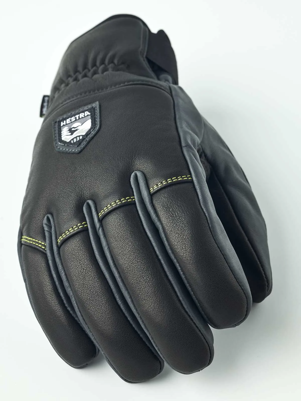 Vista dettagliata del guanto Hestra Omni Trigger in pelle nera con patch logo e cuciture gialle.