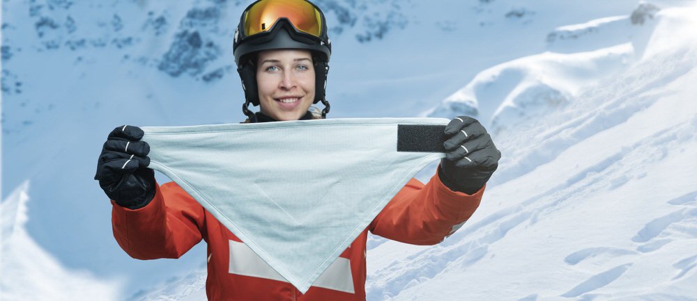 Frau mit Helm und Skibrille zeigt eine pflaumenfarbene HeiQ Filter Bandana goSnow vor verschneiter Bergkulisse.