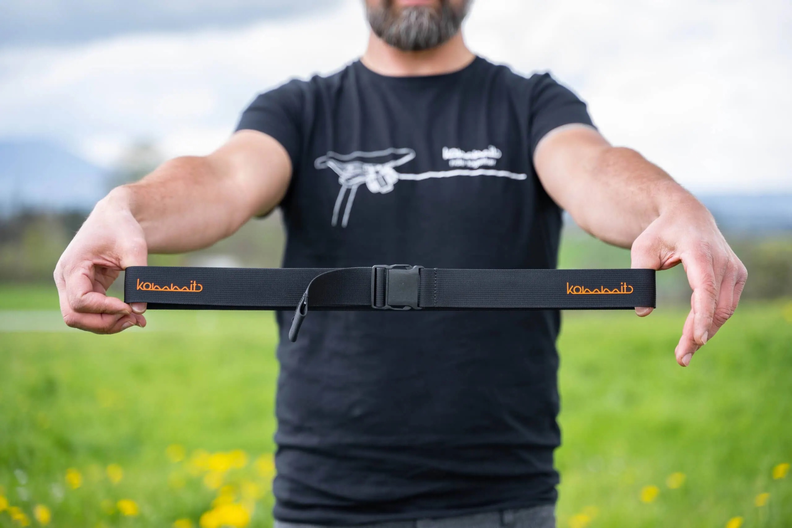 Eine Person hält einen schwarzen Outdoor-Gürtel mit orangefarbenem "kommit"-Schriftzug vor einem grünen Hintergrund.