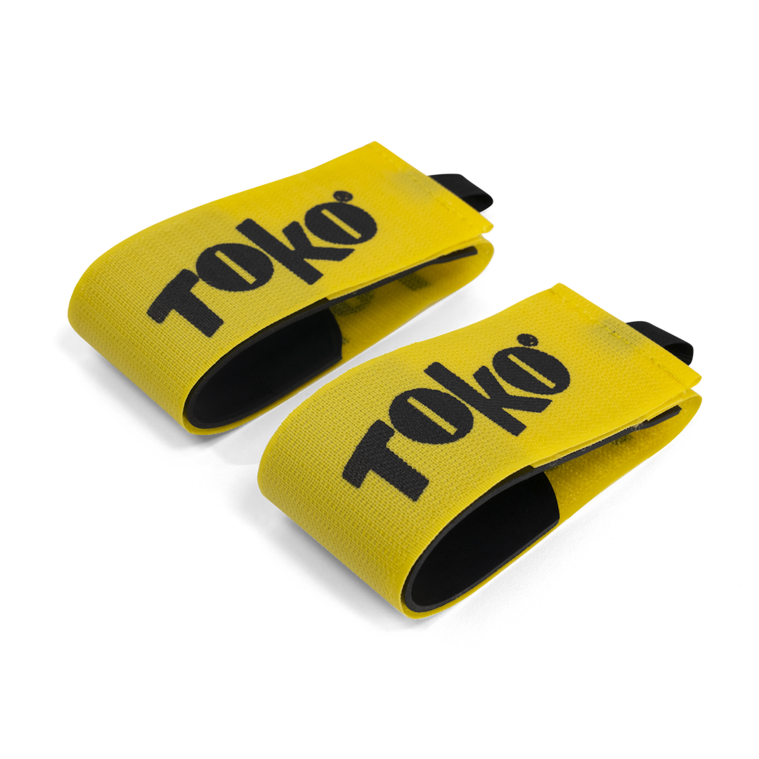 Zwei gelbe Toko Ski-Clips mit schwarzer Schrift und schwarzem Innenfutter liegen auf weißem Grund.