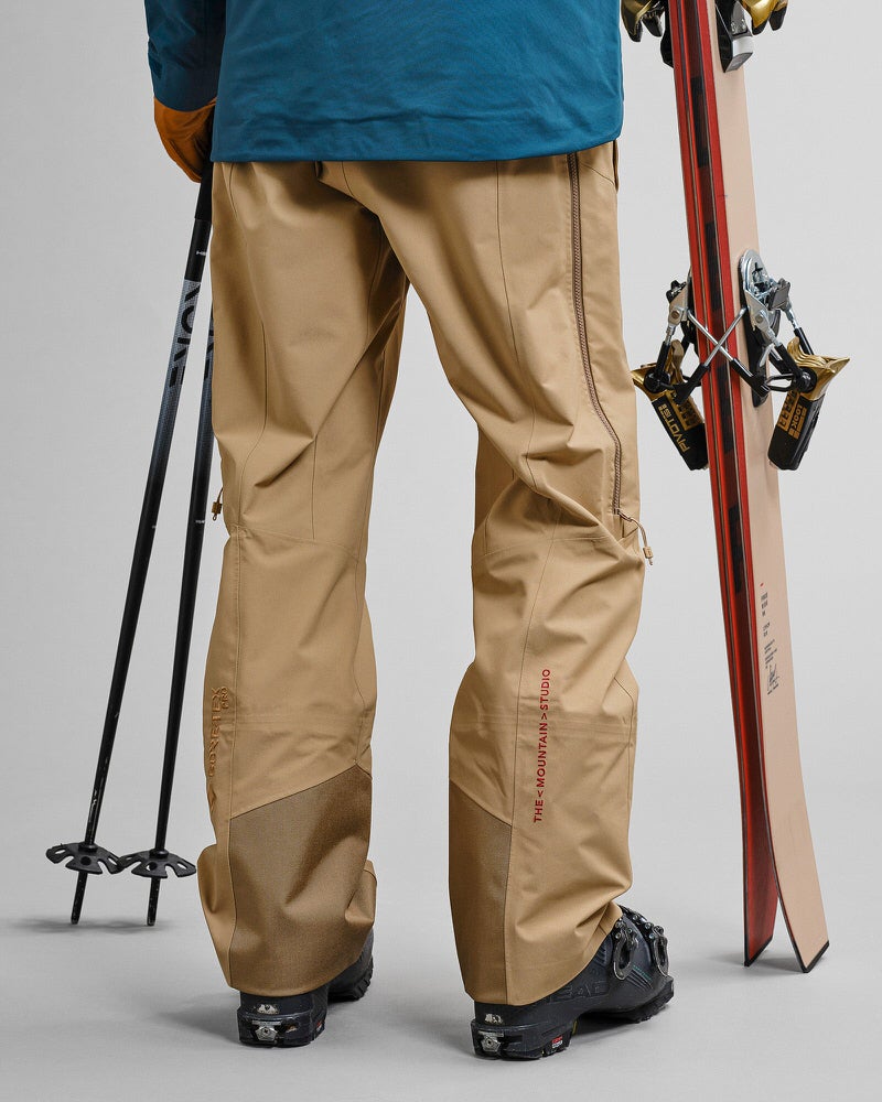 GORE-TEX Pro Skihose Person von hinten in Sandfarbener GORE-TEX Pro 3L Shell Pant, mit Ski und Skistöcken.