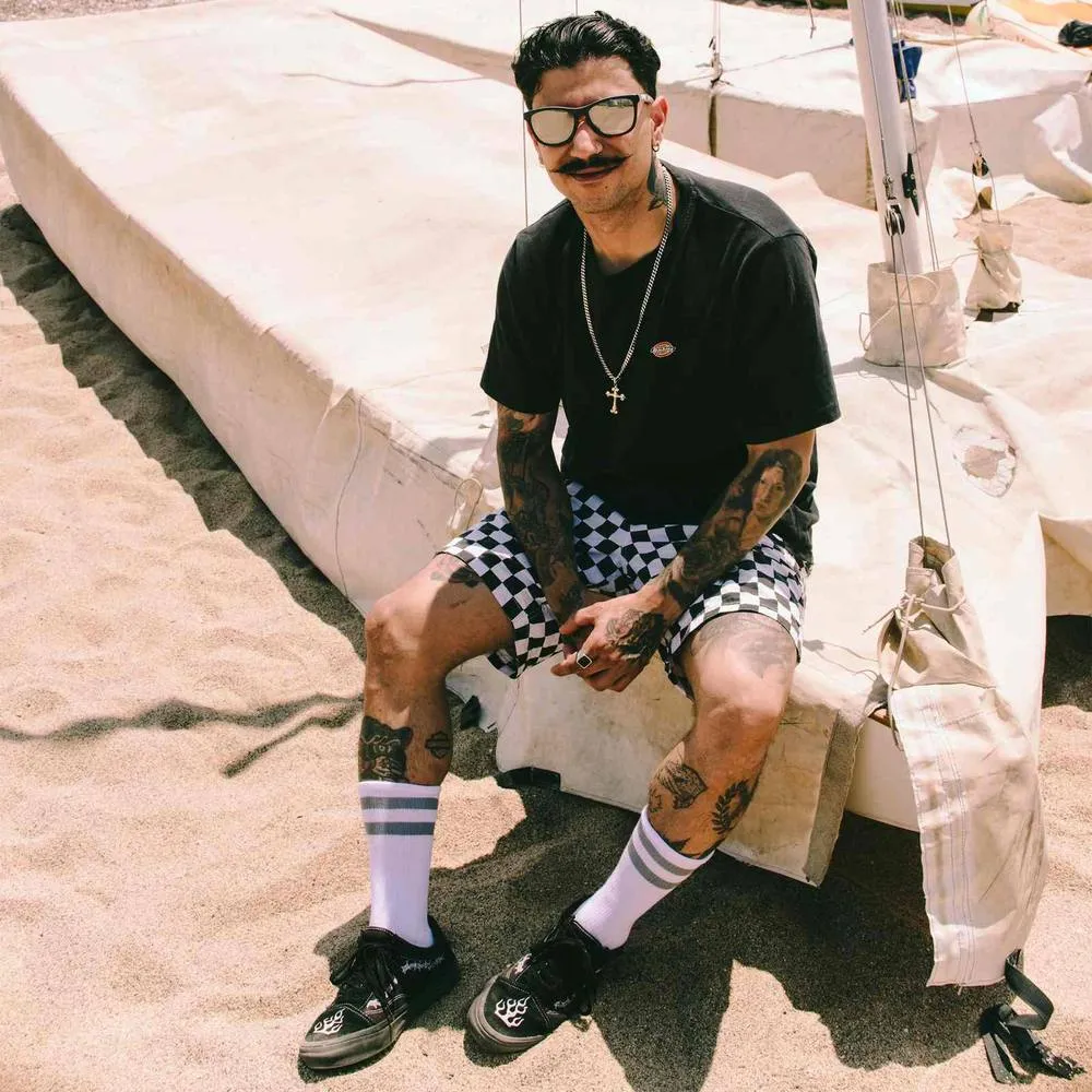 Mann mit Tätowierungen, Mid-High Socken und Schachbrettmuster-Shorts sitzt auf einer Plane am Strand.