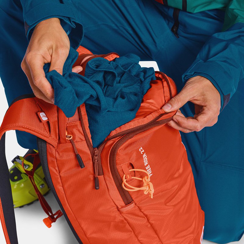 Orangefarbener Ortovox Freerider 22 Rucksack wird von einer Person mit einem blauen Kleidungsstück befüllt.