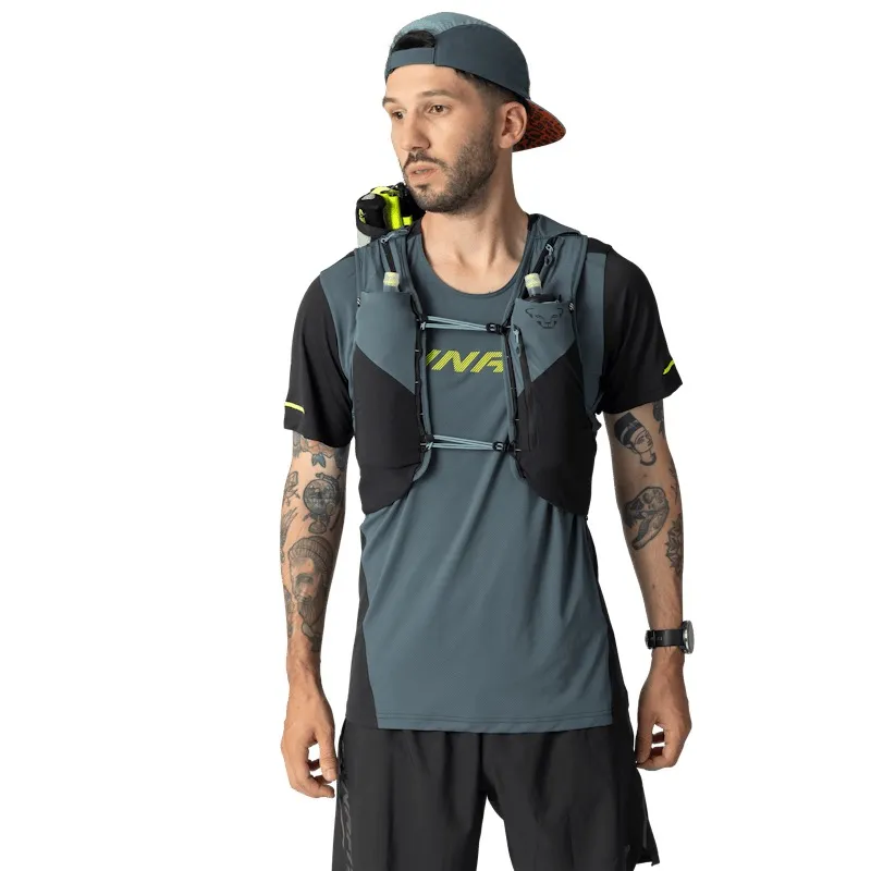 Man in athletic wear with tattoos wearing a running vest. Gray cap, looking to the left. Gray-and-black vest with pockets for water bottles. Gray T-shirt with yellow "Dynafit" print. Watch on the left. White background.