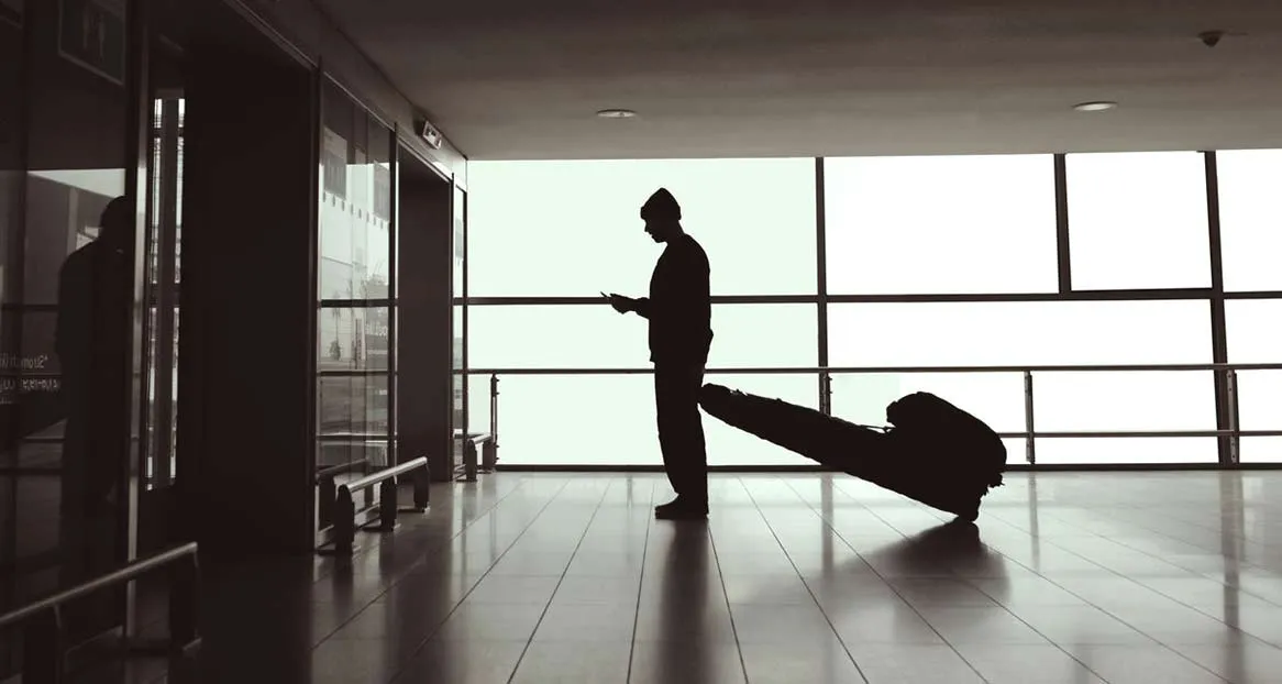 Silhouette einer Person mit Mütze, die auf ihr Handy schaut, während sie eine Snowboardtasche durch einen hellen Gang zieht.