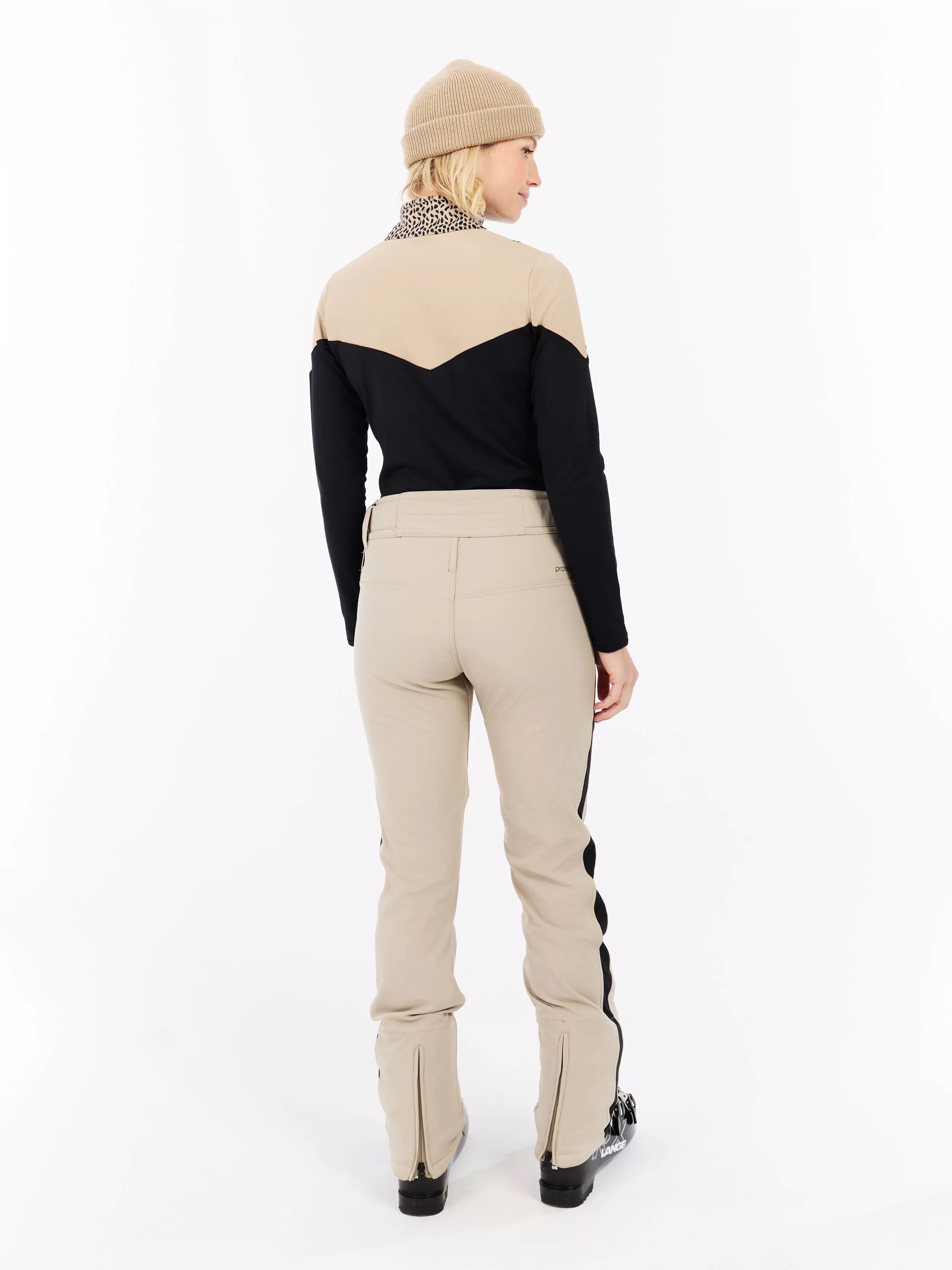 A woman with a cap, beige top and black sleeves, beige pants with black stripes and ski boots stands on a white background.