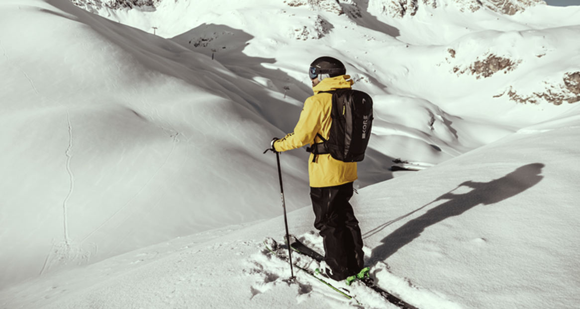 Un skieur portant une veste jaune et un sac à dos se tient dans la neige, face à des montagnes enneigées.