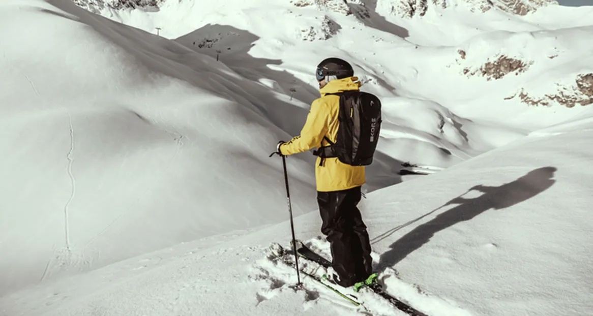 A skier with a yellow jacket and rucksack stands in the snow with a view of snow-covered mountains.