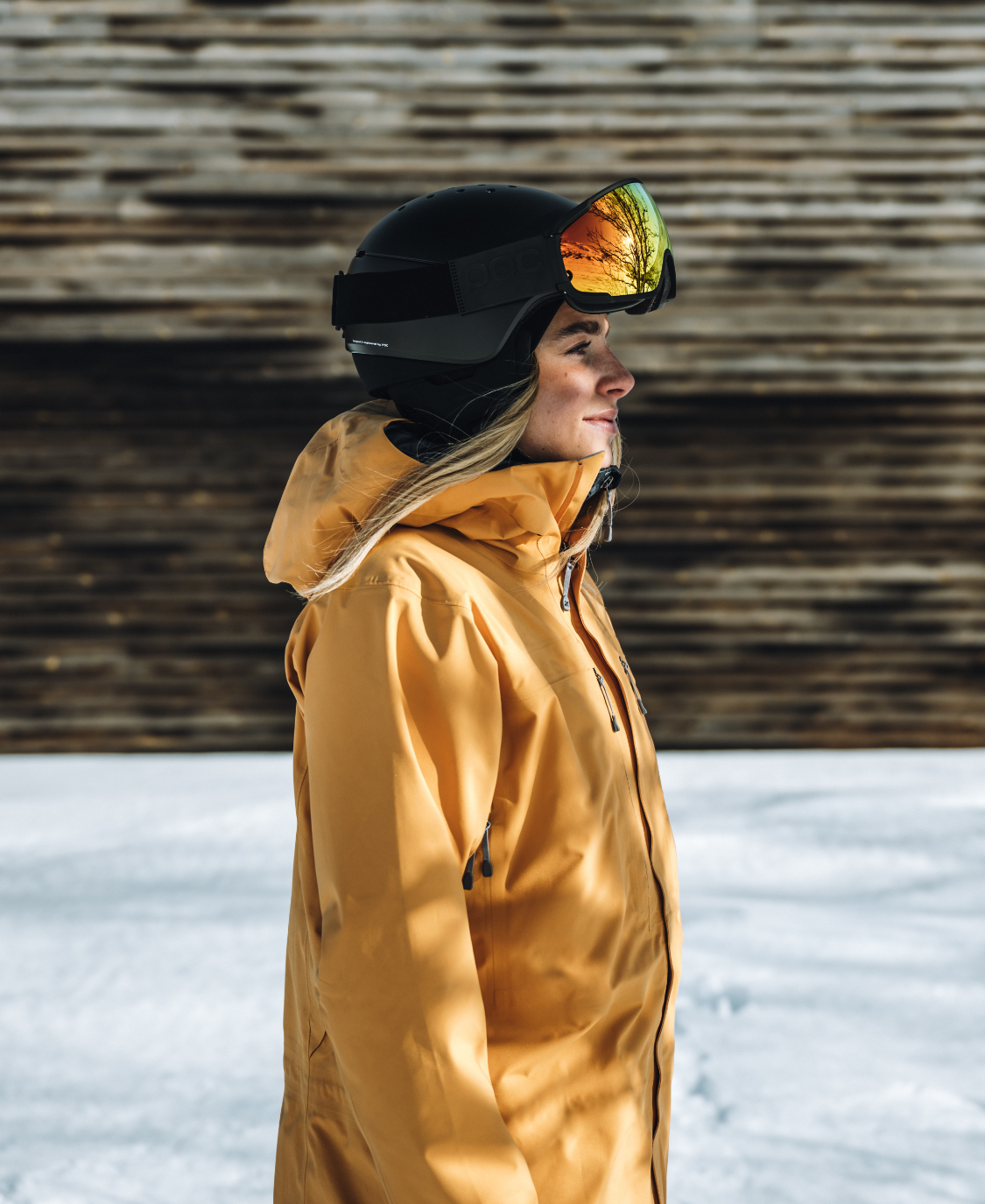 Vue de profil d'une personne portant un casque de ski POC Meninx noir et une veste jaune dans la neige. Les lunettes de ski reflètent des arbres.