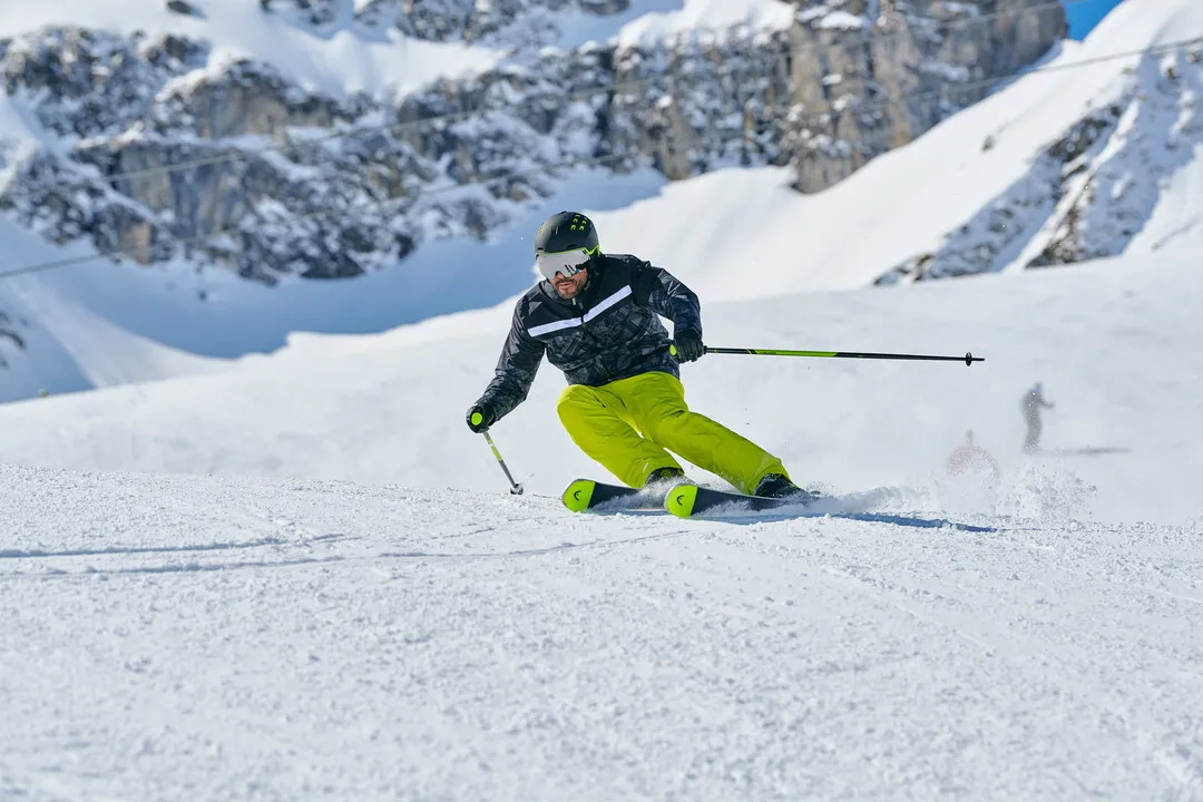 Skifahrer in grüner Hose fährt einen verschneiten Hang hinunter, im Hintergrund Berge und etwas blauer Himmel.