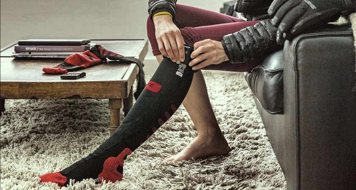 Une personne est assise sur un canapé et régule une chaussette chauffante noire à bout rouge. Des livres et une deuxième chaussette sont posés sur la table.
