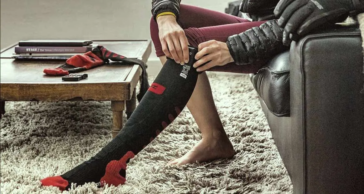 Person sitzt auf Couch und reguliert eine beheizbare, schwarze Socke mit roter Spitze. Auf dem Tisch liegen Bücher und eine zweite Socke.