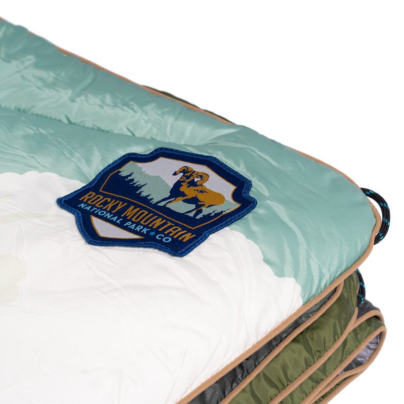 Detailansicht einer Rocky Mountain Puffy Blanket mit Logo-Patch, das ein Bergschaf zeigt. Die Decke ist in Weiß, Türkis und Olivgrün gehalten.