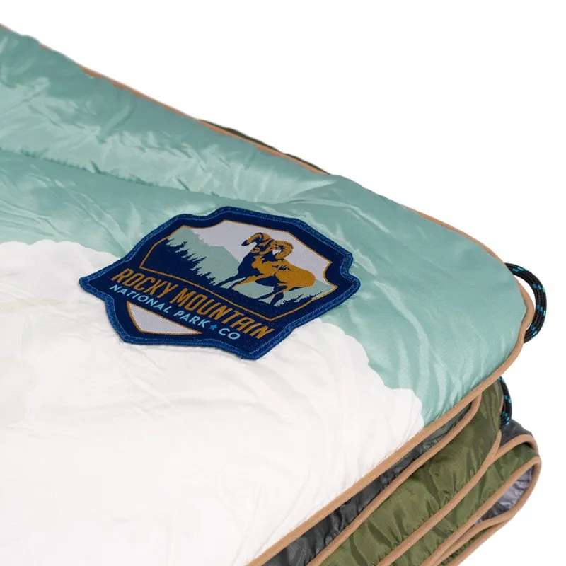 Detailansicht einer Rocky Mountain Puffy Blanket mit Logo-Patch, das ein Bergschaf zeigt. Die Decke ist in Weiß, Türkis und Olivgrün gehalten.