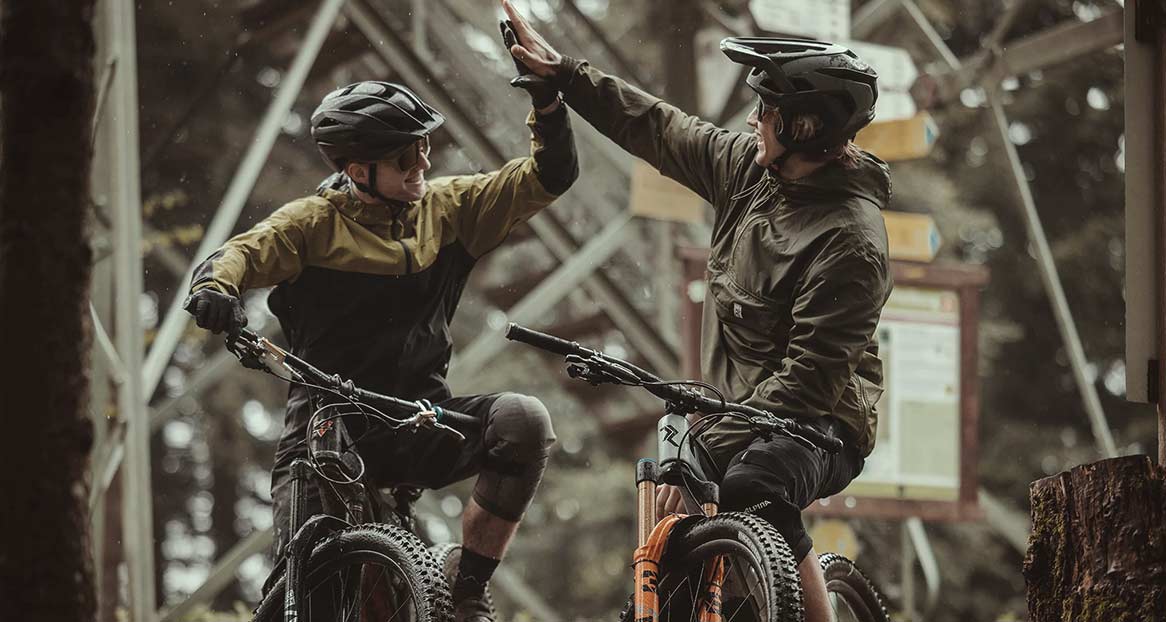 Zwei Mountainbiker in voller Ausrüstung geben sich ein High-Five vor einem unscharfen Waldhintergrund und einem hölzernen Wegweiser.
