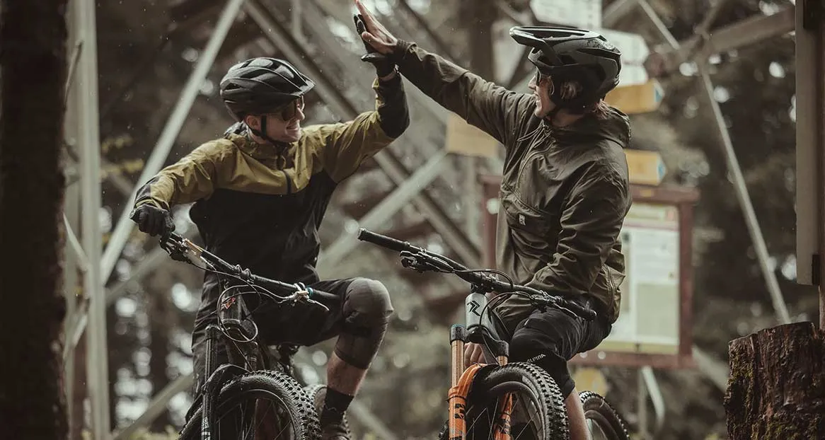 Zwei Mountainbiker in voller Ausrüstung geben sich ein High-Five vor einem unscharfen Waldhintergrund und einem hölzernen Wegweiser.